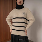 A person wearing a black and beige cable knit sweater with a round neckline, featuring horizontal stripes and a decorative floral design on the chest from BWBYAZ.