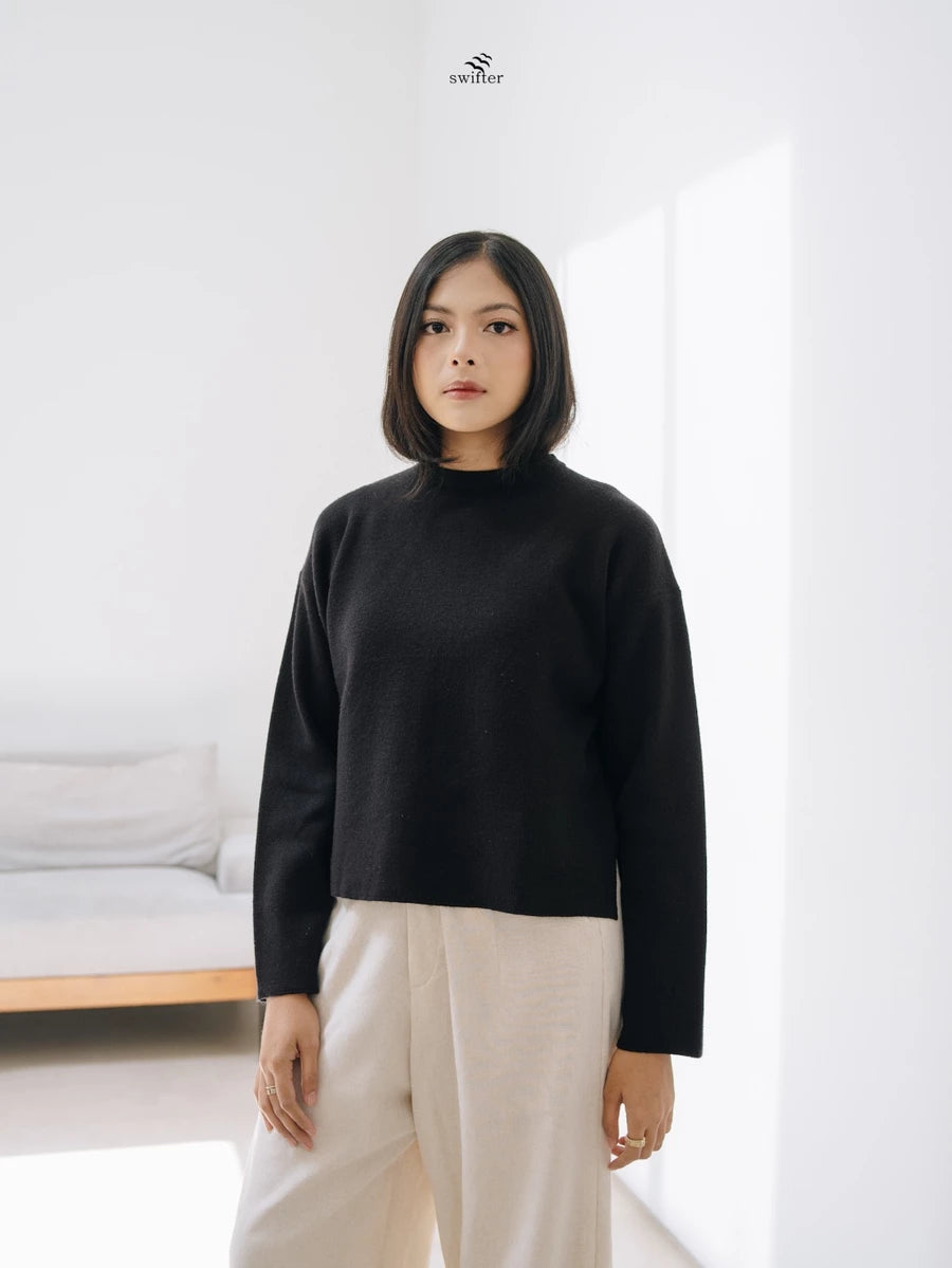 Woman wearing a black knit sweater by Swifter and beige pants in a minimalistic room.
