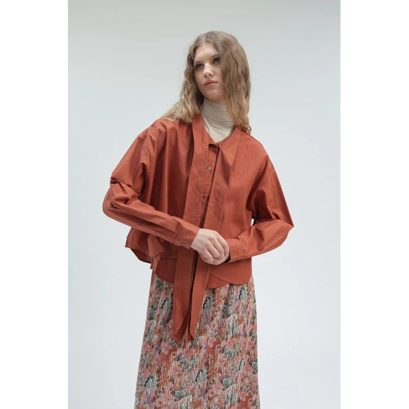 Woman wearing a terracotta women’s tie blouse by Yestoday with a loose silhouette and front button details, paired with a cream turtleneck inner and a floral pleated skirt for a modern feminine look.