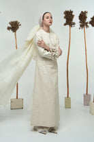 A model wearing a long white linen dress with long sleeves by Glashka, standing against a backdrop with decorative trees.