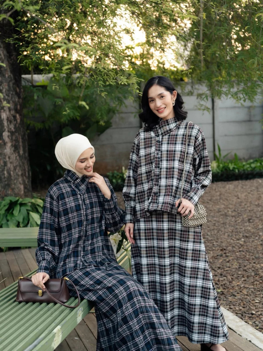 Two women posing outdoors in matching long-sleeve plaid outfits by Haidee Orlin. One is seated on a green bench wearing a white hijab and holding a brown handbag, while the other stands beside her in a dark plaid long-sleeve top and skirt set.