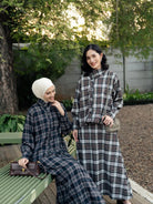 Two women posing outdoors in matching long-sleeve plaid outfits by Haidee Orlin. One is seated on a green bench wearing a white hijab and holding a brown handbag, while the other stands beside her in a dark plaid long-sleeve top and skirt set.