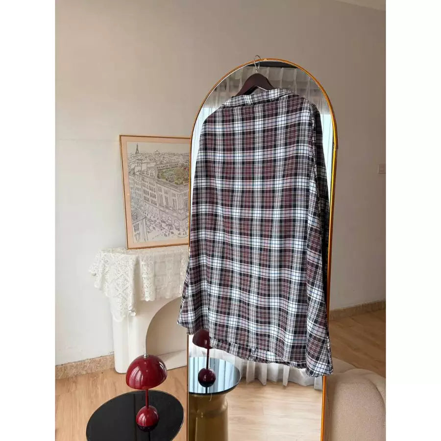 A long-sleeve plaid shirt in black, white, and red by Haidee Orlin is displayed on a hanger inside a gold-framed arched mirror, set against a warm beige wall with a white fireplace mantel in the background.