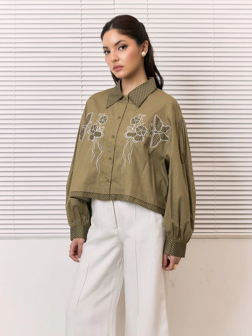 A woman models a cropped, long-sleeve button-up shirt in olive green by Studio Tui with white floral embroidery on the front, paired with white wide-leg pants, set against a background of white window blinds.