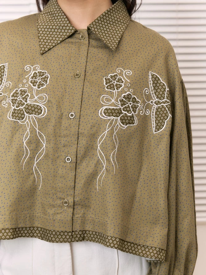 An olive green cropped button-up shirt by Studio Tui with a fine blue dot pattern, featuring white embroidered floral and butterfly designs on the front, worn by a model.