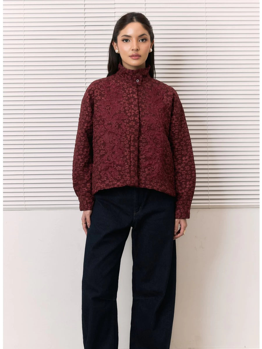 A woman stands in front of white horizontal blinds wearing a cropped, long-sleeve high-neck shirt by Studio Tui in a dark burgundy floral jacquard pattern, paired with dark navy wide-leg pants.