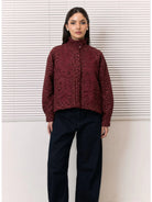 A woman stands in front of white horizontal blinds wearing a cropped, long-sleeve high-neck shirt by Studio Tui in a dark burgundy floral jacquard pattern, paired with dark navy wide-leg pants.