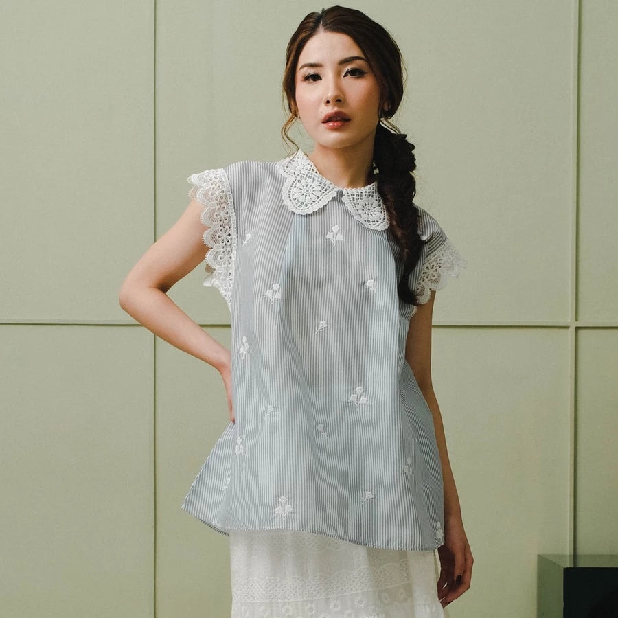 Woman wearing a light blue blouse by Holy Peony with white lace details against a green wall.