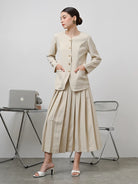 A model wearing a beige Zora skirt by Kasa Heritage with pleats, paired with a matching brown top. The skirt reaches mid-calf and has a fitted waist with a zipper at the back.