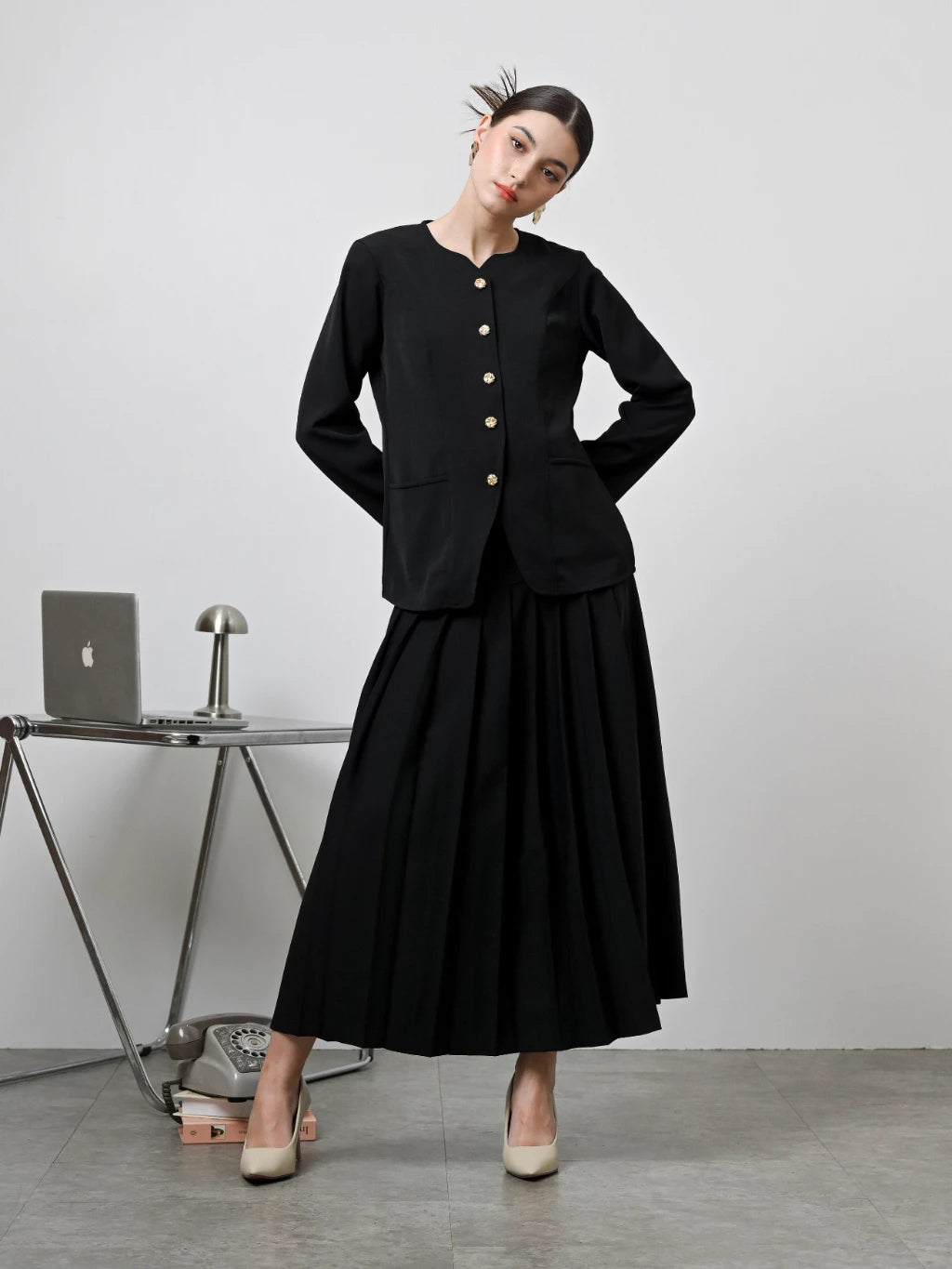 A model wearing a black Zora skirt by Kasa Heritage with pleats, paired with a matching brown top. The skirt reaches mid-calf and has a fitted waist with a zipper at the back.