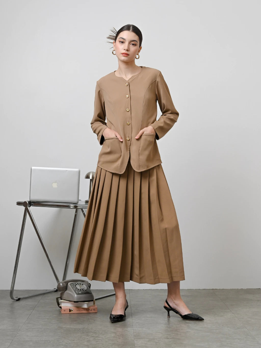 A model wearing a brown Zora skirt by Kasa Heritage with pleats, paired with a matching brown top. The skirt reaches mid-calf and has a fitted waist with a zipper at the back.